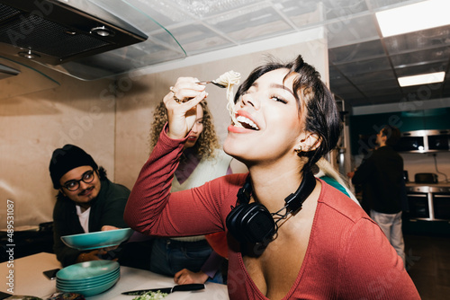 Happy young woman eating noodles by friend in kitchen at college dorm photo