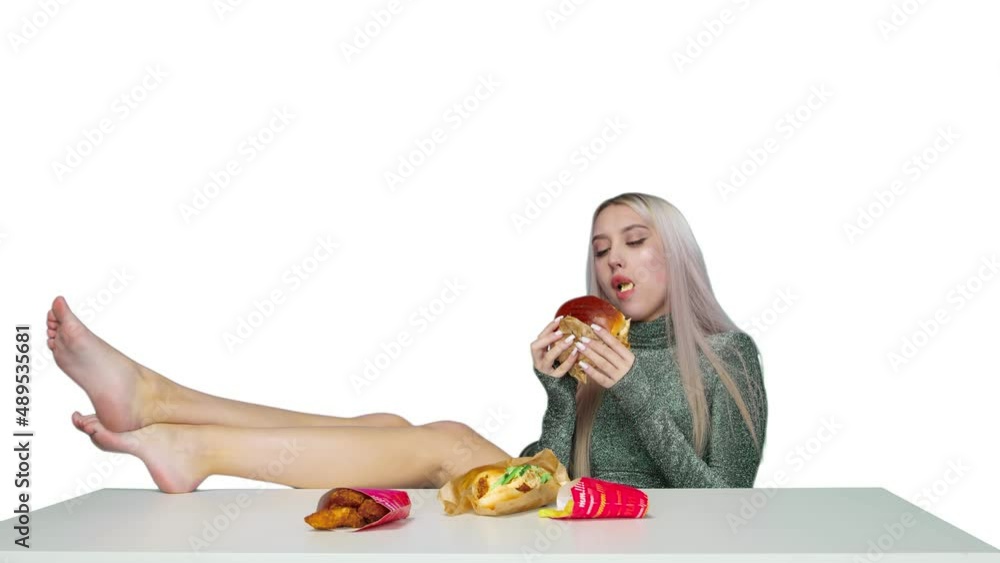 A cute girl sits on a chair with her legs on the table and eats a hamburger on a white background. Diet. The concept of healthy and unhealthy food. fast food A cute girl sits on a chair with her legs on the table and eats a hamburger on a white background. Diet. The concept of healthy and unhealthy food. fast food