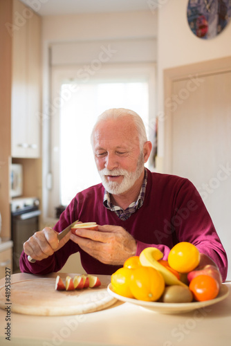 Senior man eating apple and other fruits in the kitchen on a sunny morning.