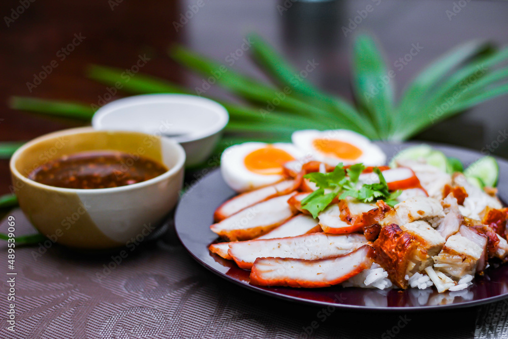 Steamed rice and red pork, crispy pork in a black plate Served with dipping sauce and dark soy sauce Place it on a black wooden table.