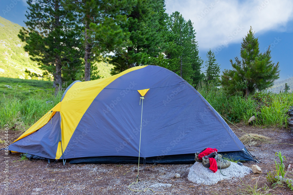 Summer camping in a tent for two on the hillside under the cedar trees. Sneakers with red socks stand on a stone near the grey and yellow tent. Active lifestyle and hiking sport.