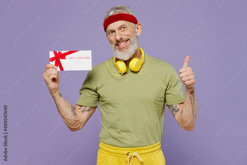 Elderly gray-haired man 40s years old in headband khaki t-shirt hold gift certificate coupon voucher card for store show thumb up like isolated on plain pastel light purple background studio portrait. Elderly gray-haired man 40s years old in headband khaki t-shirt hold gift certificate coupon voucher card for store show thumb up like isolated on plain pastel light purple background studio portrait.
