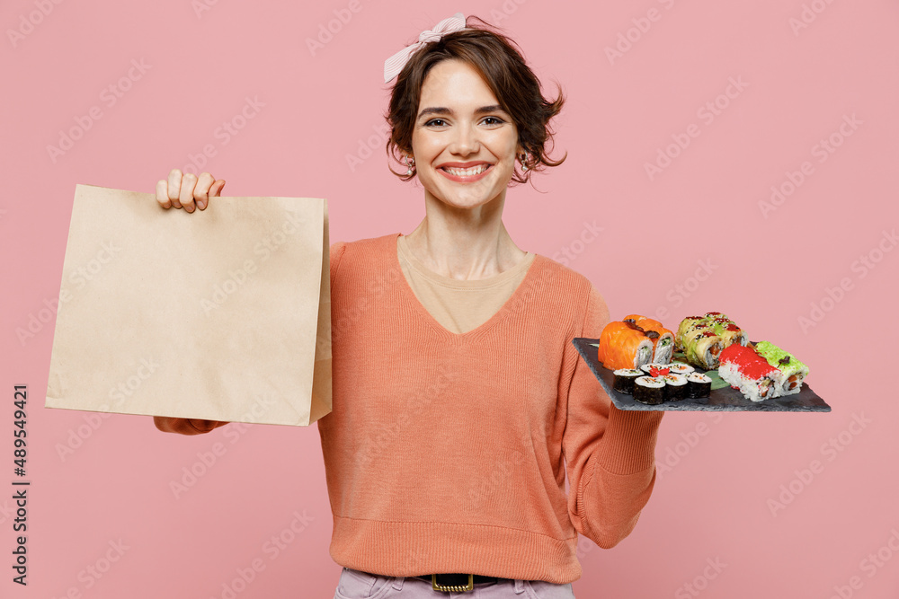 Young smiling woman in sweater hold makizushi sushi roll served on black plate traditional japanese food brown clear blank craft paper takeaway bag mock up isolated on plain pastel pink background Young smiling woman in sweater hold makizushi sushi roll served on black plate traditional japanese food brown clear blank craft paper takeaway bag mock up isolated on plain pastel pink background
