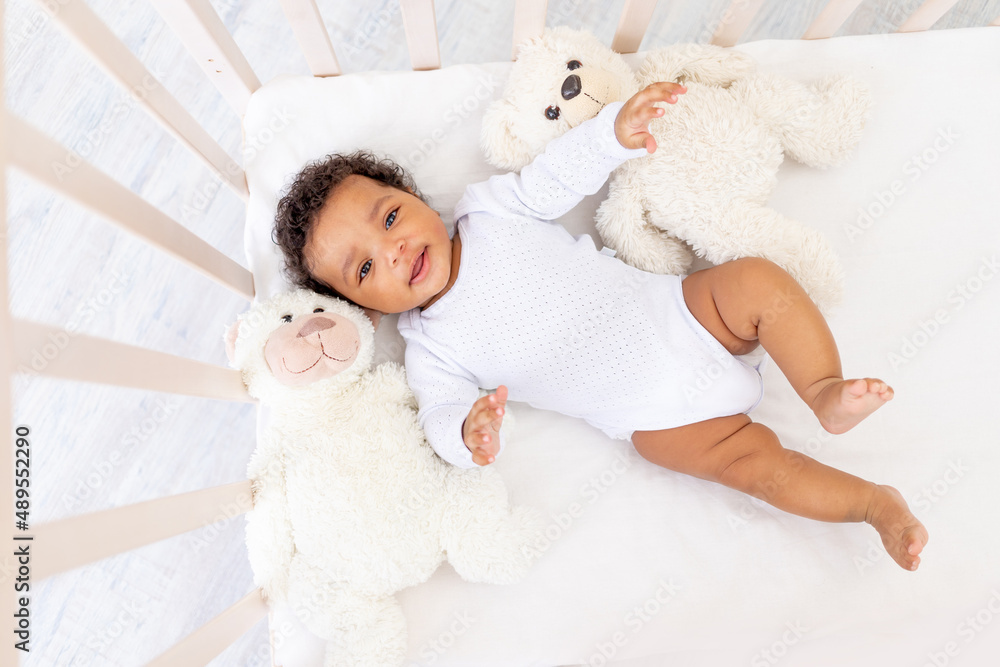 Smiling African-American little baby in white sleeping bed with bear toys