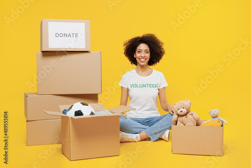 Full body smiling young woman of African American ethnicity wear white volunteer t-shirt sit near boxes with presents isolated on plain yellow background. Voluntary free work assistance help concept photo