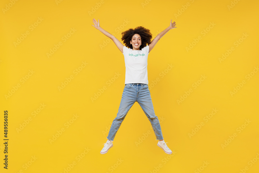 Full body young woman of African American ethnicity in white volunteer t-shirt jump high with outstretched hands isolated on plain yellow background. Voluntary free work assistance help grace concept. Full body young woman of African American ethnicity in white volunteer t-shirt jump high with outstretched hands isolated on plain yellow background. Voluntary free work assistance help grace concept.