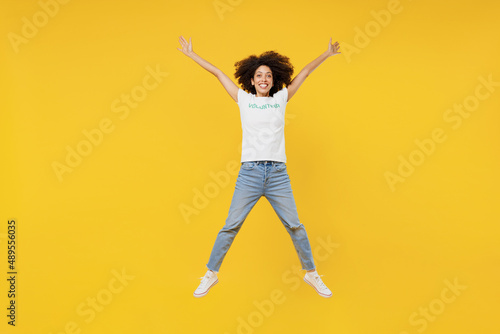 Full body young woman of African American ethnicity in white volunteer t-shirt jump high with outstretched hands isolated on plain yellow background. Voluntary free work assistance help grace concept.