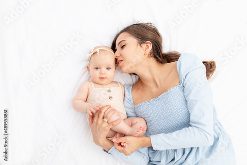 mom with a newborn baby girl in a cotton suit on the bed at home hugging and kissing him, happy motherhood or family