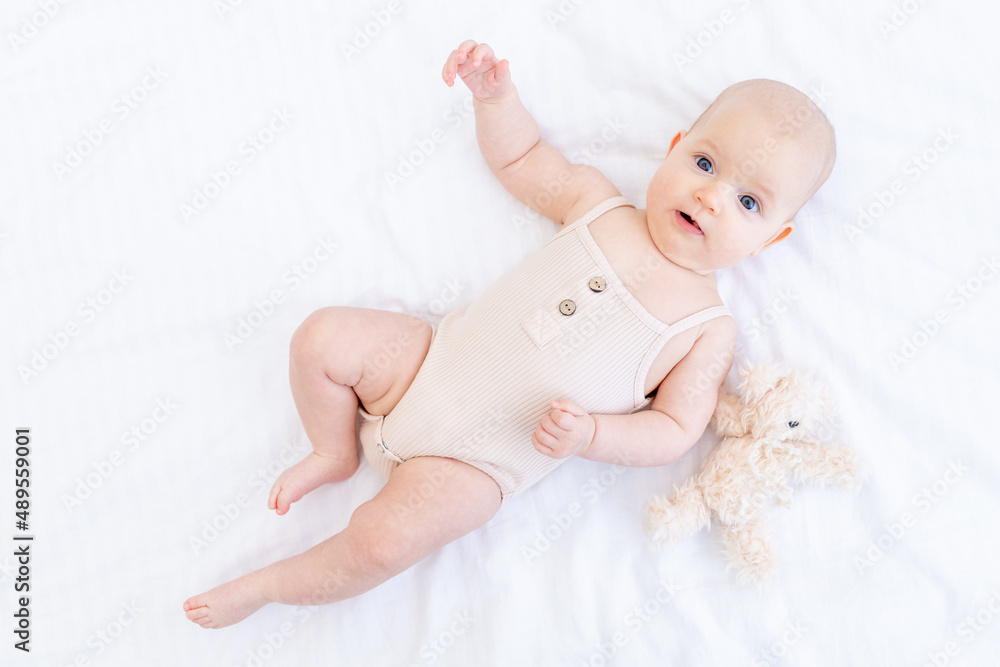a newborn baby baby girl in a cotton suit on a white bed at home is lying and smiling with a teddy bear a newborn baby baby girl in a cotton suit on a white bed at home is lying and smiling with a teddy bear