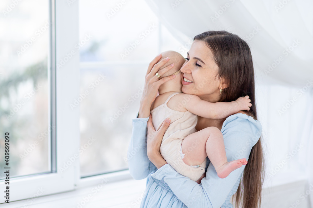 mom holds a newborn baby girl in a cotton suit at the window of the house hugging and kissing him, happy motherhood or family mom holds a newborn baby girl in a cotton suit at the window of the house hugging and kissing him, happy motherhood or family