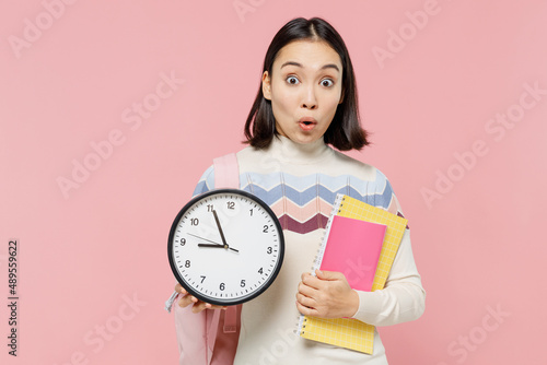 Shocked happy teen student girl of Asian ethnicity wear sweater backpack hold books look at clock isolated on pastel plain light pink background Education in high school university college concept