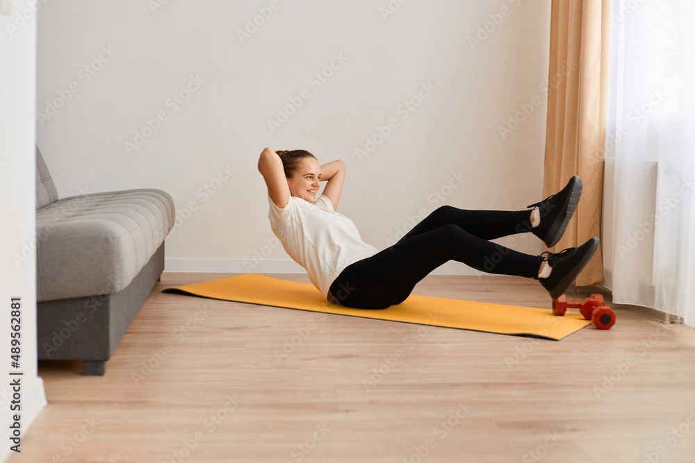 Portrait of athletic woman wearing white t shirt and black leggins doing sport exercises at home, practicing, doing crisscross exercise, bicycle crunches pose, working out.