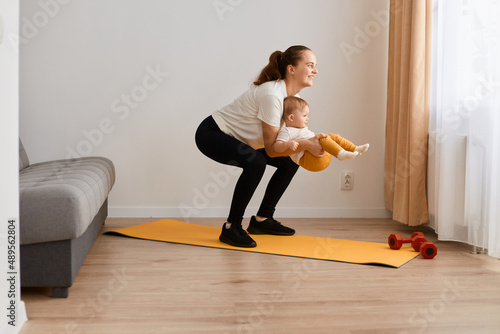 Image of attractive sporty athletic woman wearing white t shirt and black leggins doing sport exercises at home, doing squats with toddler daughter in hands.