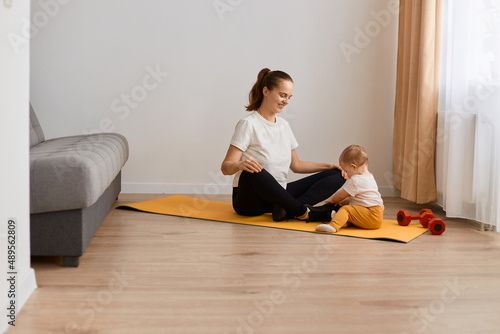 woman wearing white t shirt and black leggins doing sport exercises at home, sitting in lotus position, practicing yoga, looking at her toddler daughter with happy smile.