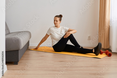 indoor shot of young adult attractive woman wearing white t shirt and black leggins doing sport exercises at home, sitting on mat and doing exercise flexibility, looking away.
