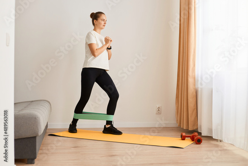 indoor shot of athletic young adult woman wearing white t shirt and black leggins doing sport exercises at home, standing on yoga mat and training legs with elastic band.