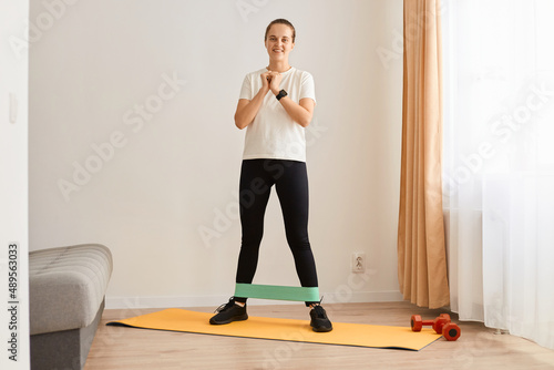 Indoor full length shot of sporty woman wearing white t shirt and black leggins doing sport exercises at home, standing and doing workout for legs, using resistance band for training.