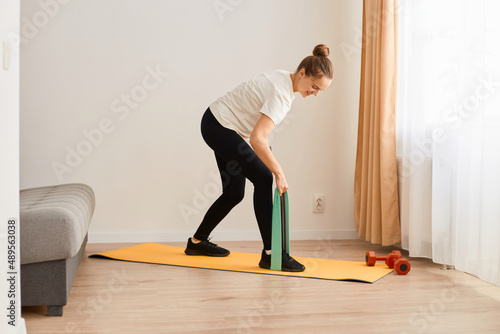 Portrait of slim attractive woman wearing white t shirt and black leggins doing sport exercises at home, standing with bending upper body and holding resistance band for workout.