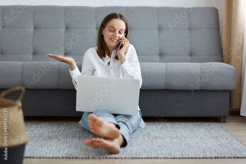 Φωτογραφία Horizontal shot of Caucasian beautiful woman wearing white shirt and jeans sitting on floor, having helpless facial expression, spreading hand aside, talking phone and using laptop