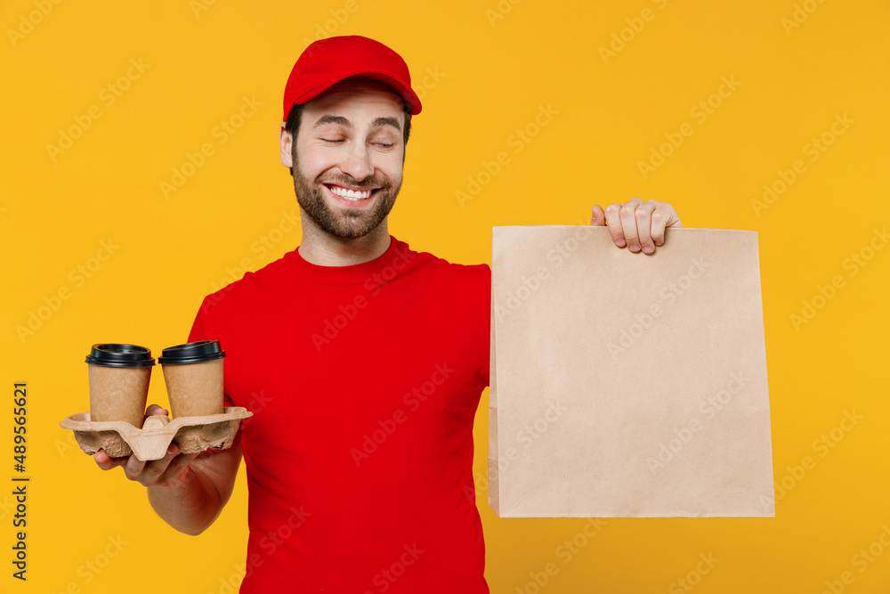 Delivery boy employee man in red cap T-shirt uniform work as dealer courier hold takeaway craft brown cup coffee to go brown clear blank craft paper takeaway bag isolated on plain yellow background. Delivery boy employee man in red cap T-shirt uniform work as dealer courier hold takeaway craft brown cup coffee to go brown clear blank craft paper takeaway bag isolated on plain yellow background.