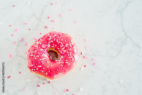A donut with pink frosting and Valentines pink, white, and red heart sprinkles on a white marble background