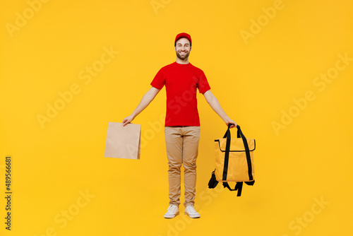 Full body delivery guy employee man in red cap T-shirt uniform work as dealer courier hold brown blank craft paper takeaway bag mock up thermal food bag backpack isolated on plain yellow background.