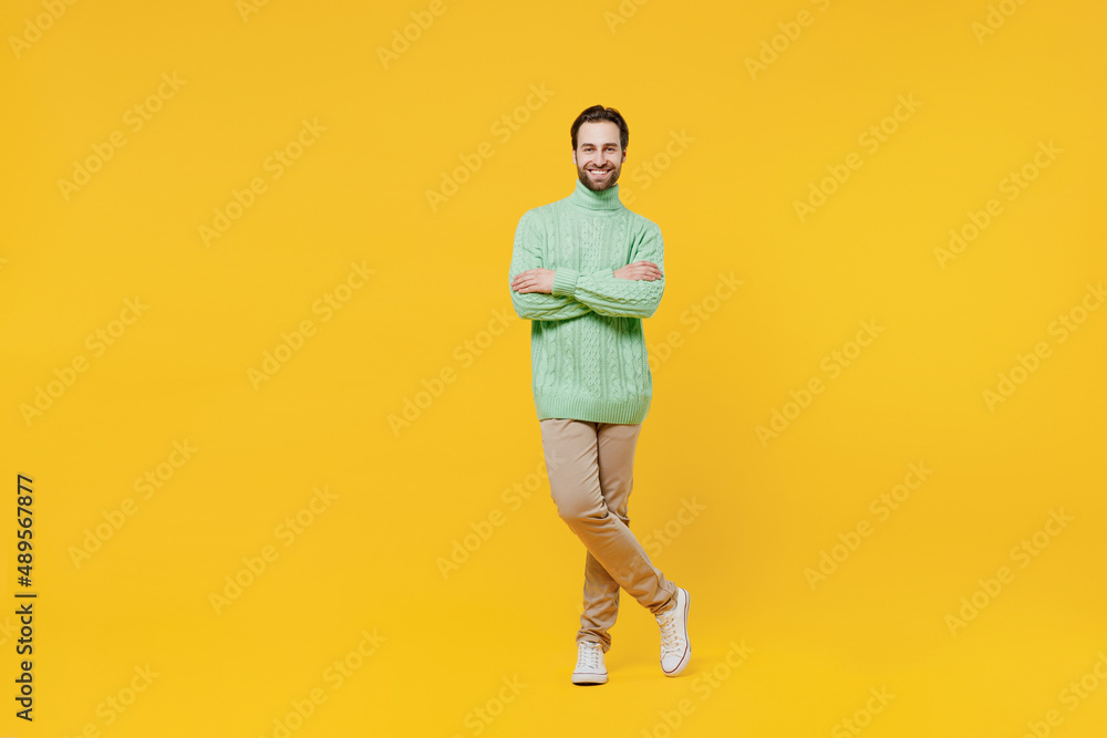 Fototapeta premium Full body smiling happy cheerful young man 20s wearing mint knitted sweater hold hands crossed folded look camera isolated on plain yellow background studio portrait. People lifestyle fashion concept.