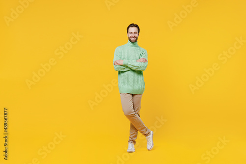 Full body smiling happy cheerful young man 20s wearing mint knitted sweater hold hands crossed folded look camera isolated on plain yellow background studio portrait. People lifestyle fashion concept.