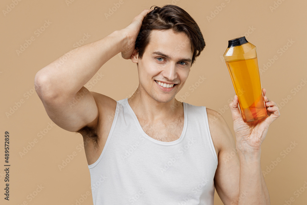Attractive smiling happy young man 20s perfect skin in undershirt hold bottle of shampoo touch hair isolated on pastel pastel beige background studio. Skin care healthcare cosmetic procedures concept. Attractive smiling happy young man 20s perfect skin in undershirt hold bottle of shampoo touch hair isolated on pastel pastel beige background studio. Skin care healthcare cosmetic procedures concept.