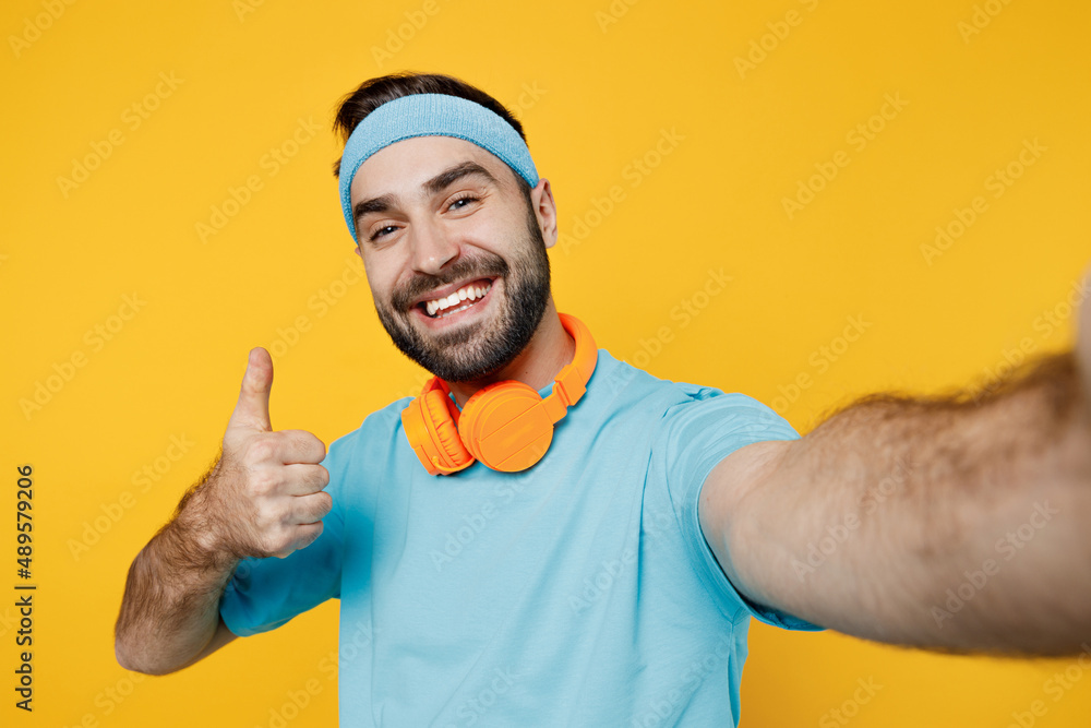 Close up young fitness trainer instructor sporty man sportsman wear headband blue t-shirt do selfie shot pov on mobile phone show thumb up isolated on plain yellow background. Workout sport concept. Close up young fitness trainer instructor sporty man sportsman wear headband blue t-shirt do selfie shot pov on mobile phone show thumb up isolated on plain yellow background. Workout sport concept.