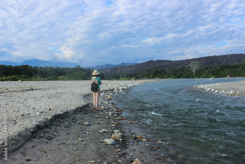 Mujer blanca en traje de baño verde con mochila negra contemplando el paisaje a la orilla del río. photo