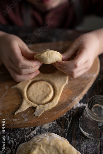 Kid making pelmeni (meet dumplings) of dough on wooden table with ingredients flour, oil, salt, dark background. Copy space. Home bakery concept, kitchen cooking story.