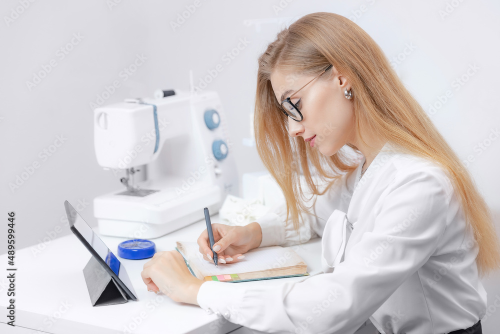 Young woman seamstress watches video lessons on tablet for sew. Concept online tailor clothes Young woman seamstress watches video lessons on tablet for sew. Concept online tailor clothes