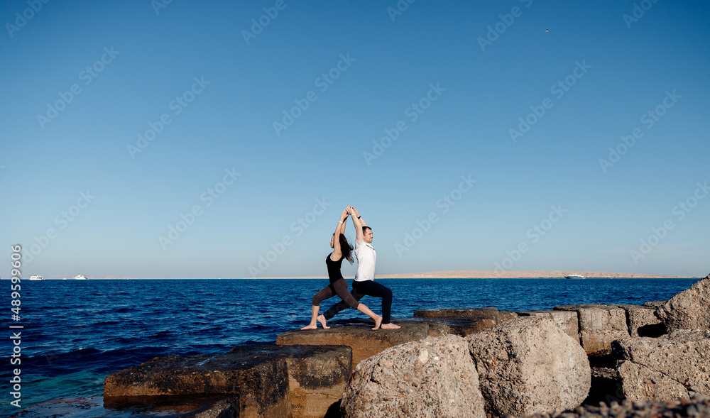 Couple sporty people practice yoga in pair, man and woman asana background blue sky and sea Couple sporty people practice yoga in pair, man and woman asana background blue sky and sea