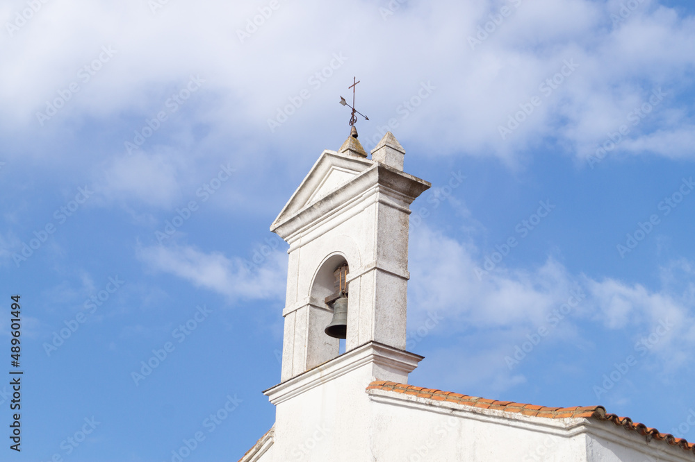 Bell tower of the chapel of San Pedro de Aracena (Huelva, Spain). White façade of a Christian place of worship with a large bell and a weathervane in the shape of an arrow and a cross.