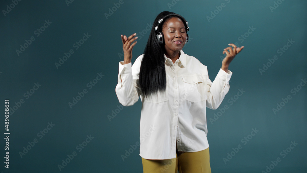 A cute charming african woman in a white blouse with earpieces is dancing on color background