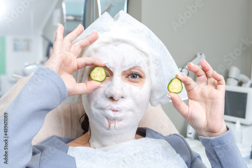 A young pretty woman with a cream mask on her face holds a plastic cucumber in her hands.