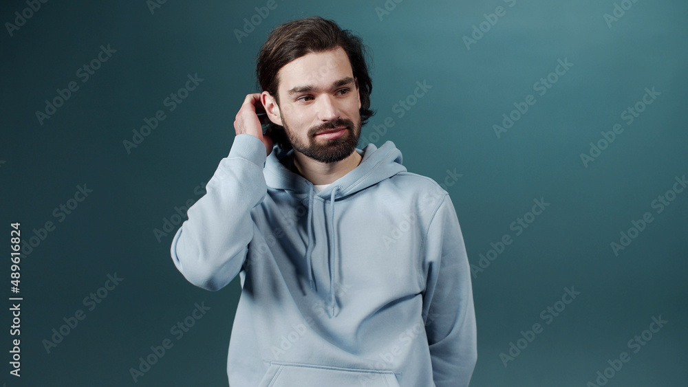 A shy bearded man is standing, fixing his hair and looking at the camera