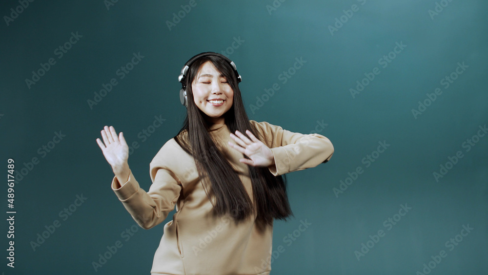 Cute charming Chinese woman in beige sweatshirt with earpieces dancing on color background