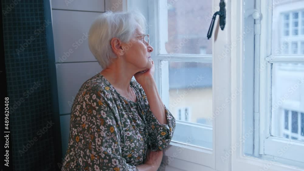 An elderly woman pensioner looks out the window sitting in her house. High quality 4k footage An elderly woman pensioner looks out the window sitting in her house. High quality 4k footage