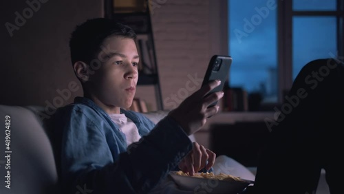 happy teen boy using phone at night at home and eating potato chips, chatting with friends, playing game indoors