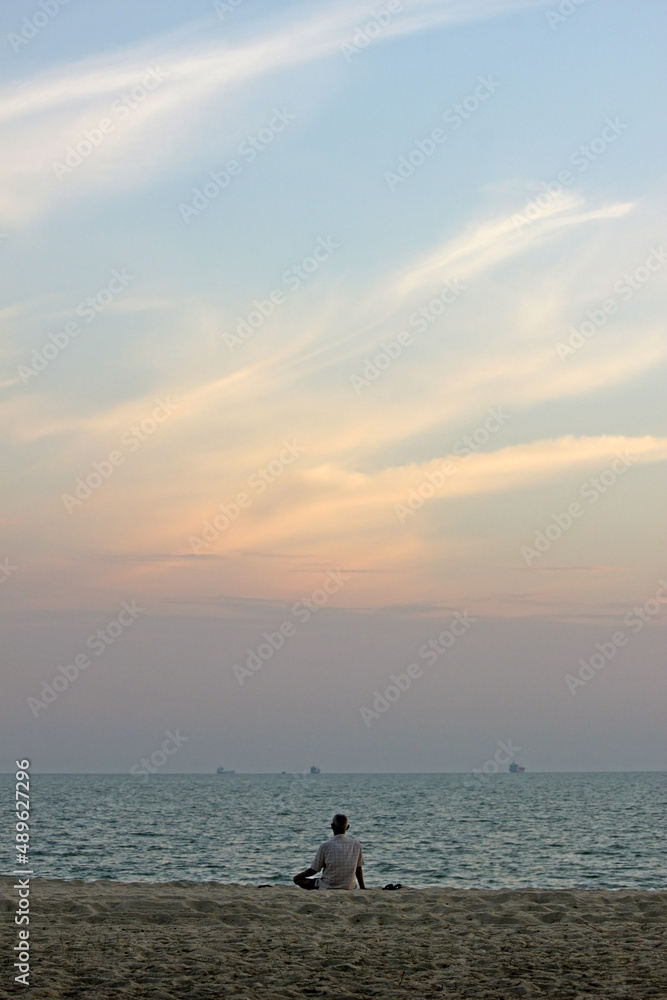 Back of a senior Asian man sitting on the sand at a beach looking at the ocean and the sunset sky, southeast Asia