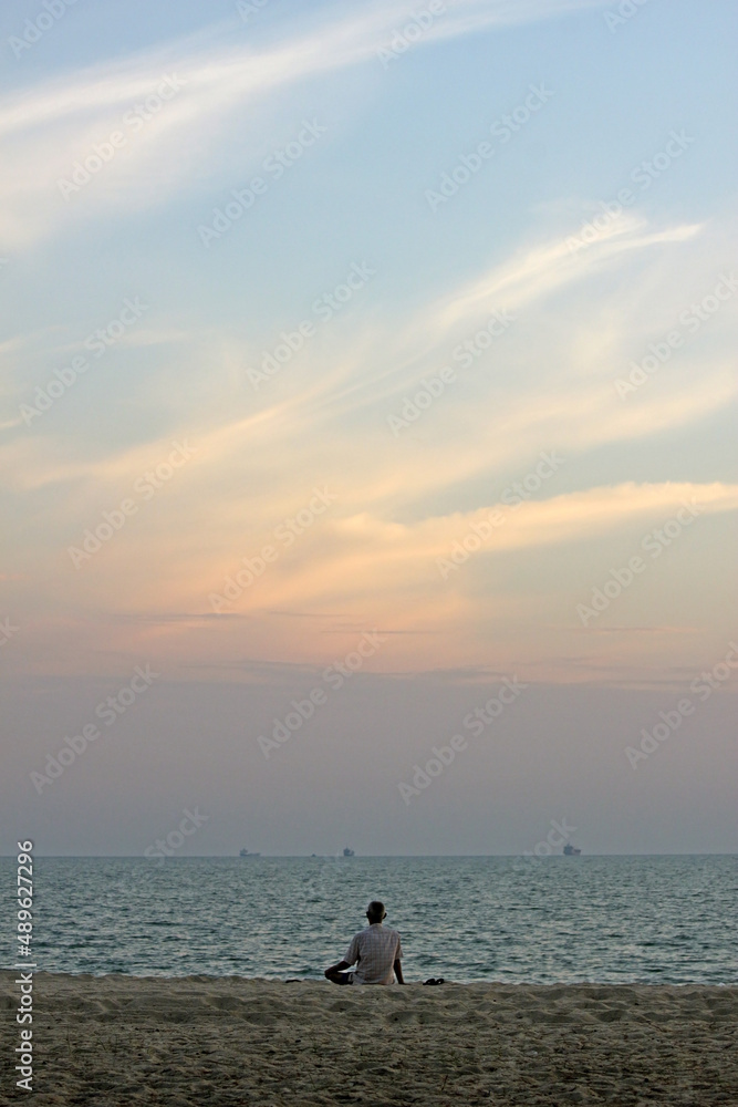 Fototapeta premium Back of a senior Asian man sitting on the sand at a beach looking at the ocean and the sunset sky, southeast Asia