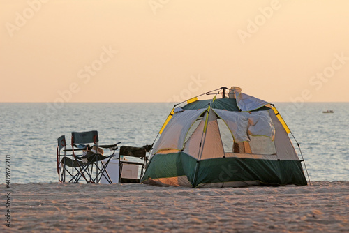 Dome tent pitched up on a beach with capming chairs next to it. Ocean view with clear orange sunset sky on the horizon. No people, eastern Thailand.