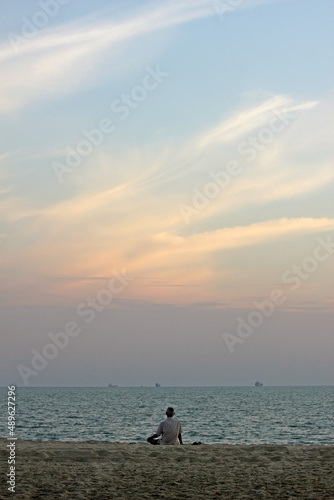 Back of a senior Asian man sitting on the sand at a beach looking at the ocean and the sunset sky, southeast Asia