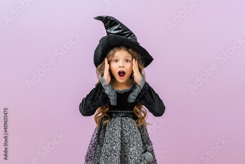 A surprised and joyful sorceress in a black fancy dress on an isolated background. A little girl in a witch costume with curly hair.