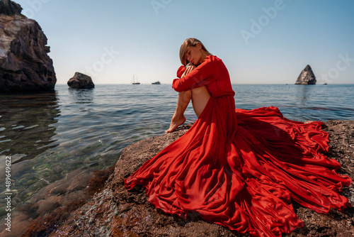 Fotografia Beautiful sensual woman in a flying red dress and long hair, sitting on a rock above the beautiful sea in a large bay