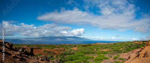 Looking across a rugged landscape  on Lanai of red dirt, growing shrubs and boulders to the ocean and the island of Maui across the channel.