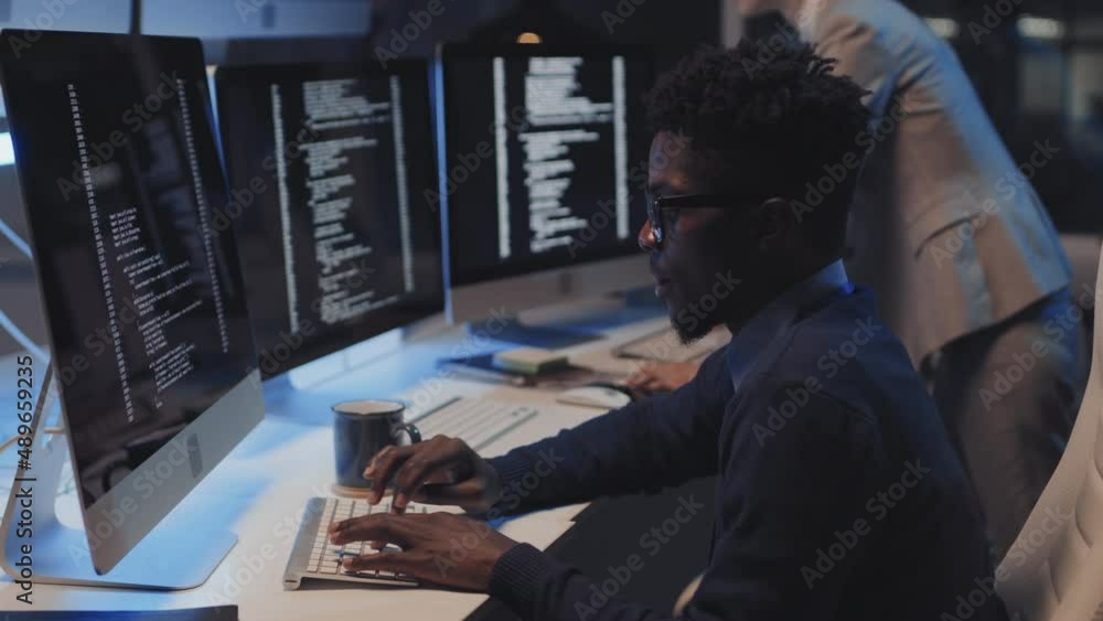 Side view of young black man wearing formal clothes and eyeglasses, sitting on chair at desk in corporate office, coding on computer at night, typing on keyboard, colleague standing on background Side view of young black man wearing formal clothes and eyeglasses, sitting on chair at desk in corporate office, coding on computer at night, typing on keyboard, colleague standing on background
