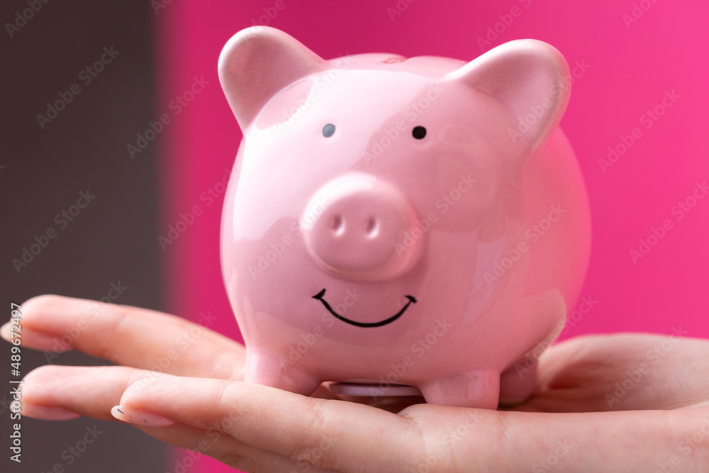 A cute girl in a pink jacket is holding a 3d piggy bank in her hands. Beautiful close-up portrait in studio on pink.